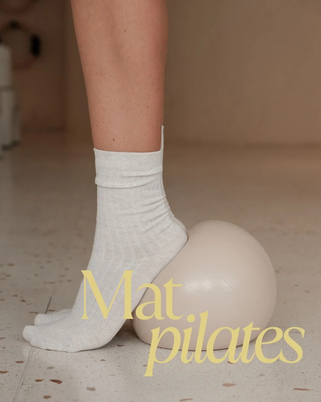 Una gamba con calzino bianco, appoggiata su una palla di Pilates bianca, sul pavimento di un locale neutro. La scritta in sovrimpressione è 'Mat, pilates'.