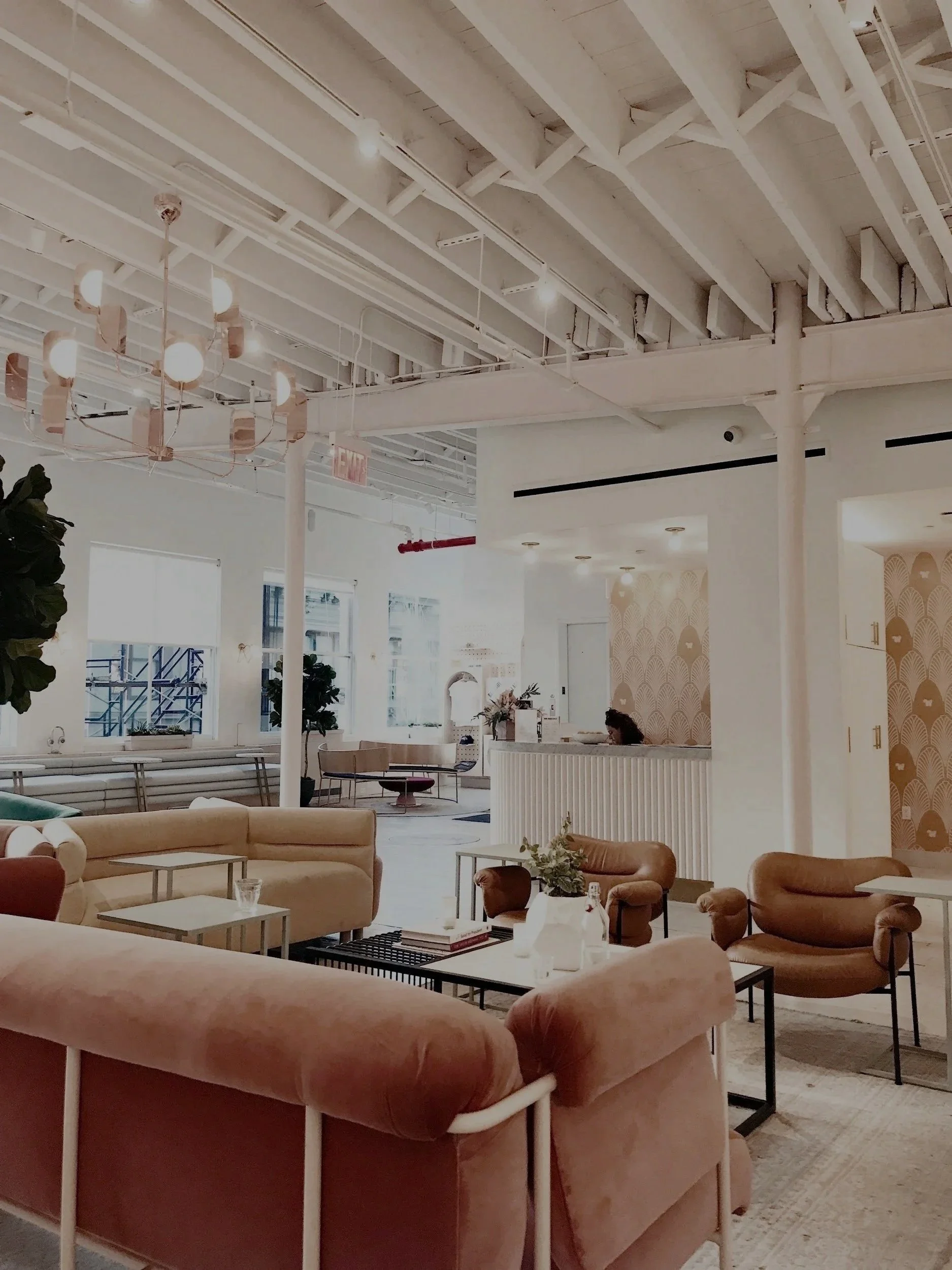 Modern, bright lounge with beige and brown sofas and chairs, white walls, high ceiling with exposed beams, decorative wallpaper, and ambient lighting.