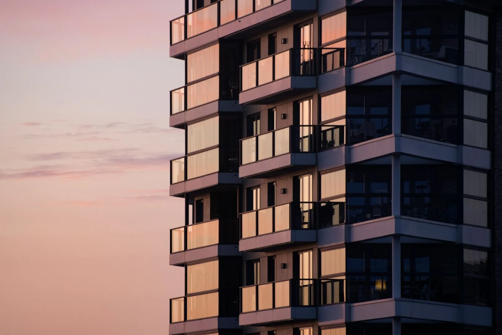 Close-up of a modern high-rise apartment building during sunset, with glass balconies reflecting the Pink and orange sky.