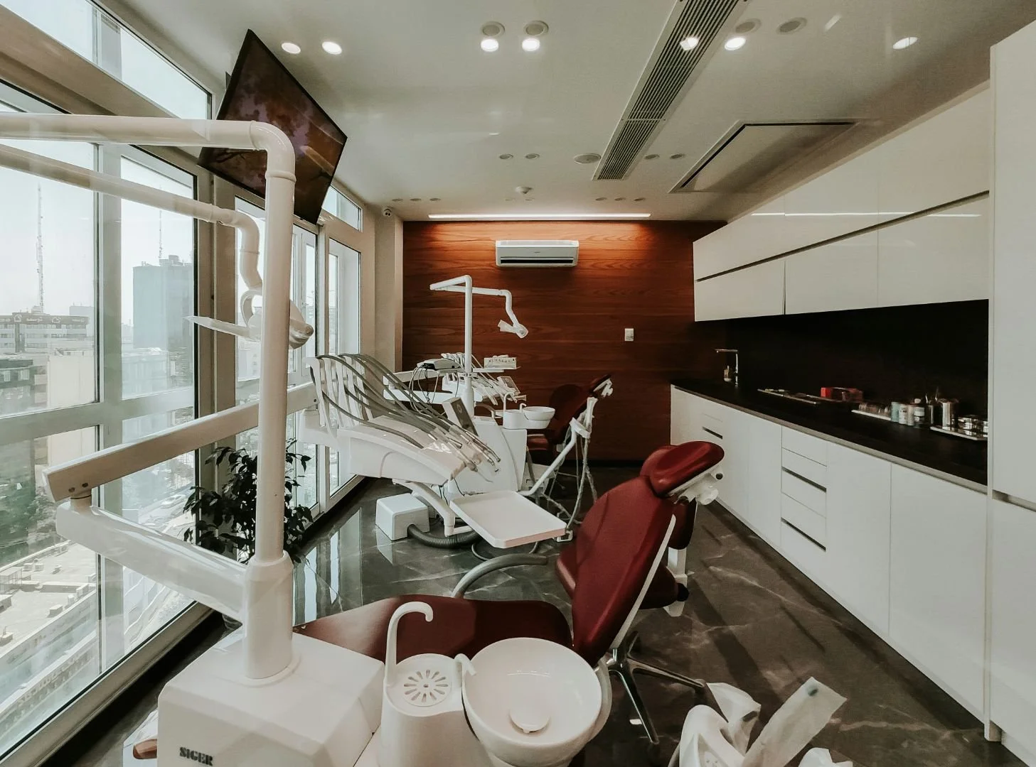 A modern dental treatment room with white cabinets, a black countertop, and red dental chairs near large windows showing a cityscape. Dental equipment and tools are present, and there is a television mounted on the wall.