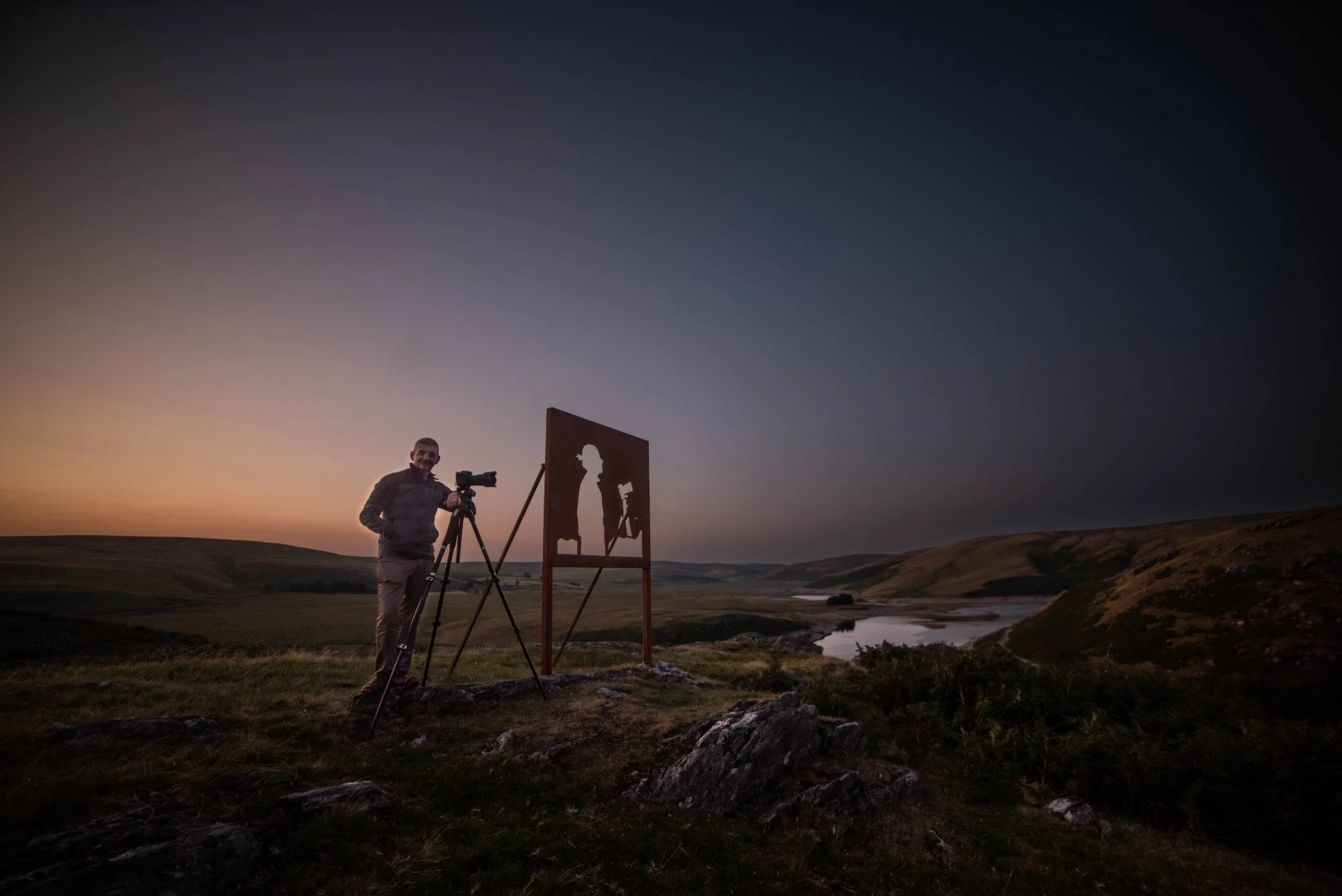 A man using a camera on a tripod setting up a shot in a natural landscape during sunset with hills, a river, and a silhouette of a painting of a person in the background. Alyn Wallace memorial in The Cambrian Mountains, Mid Wales.