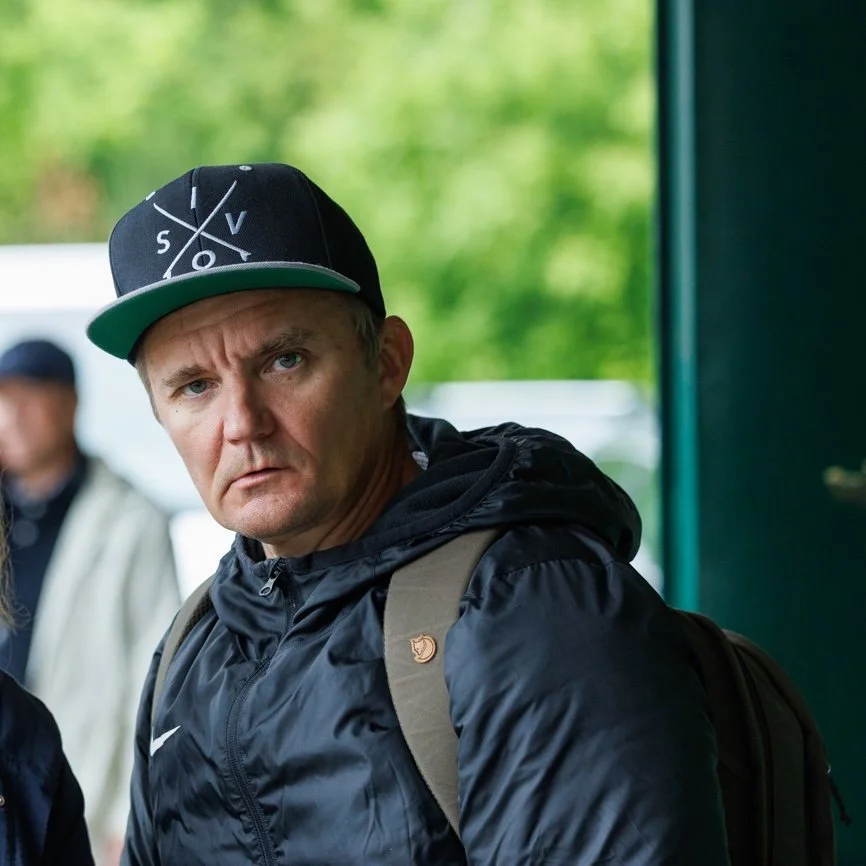 A man with a serious expression wearing a black cap with white design, a black jacket, and a backpack, standing outdoors with a blurred green background.