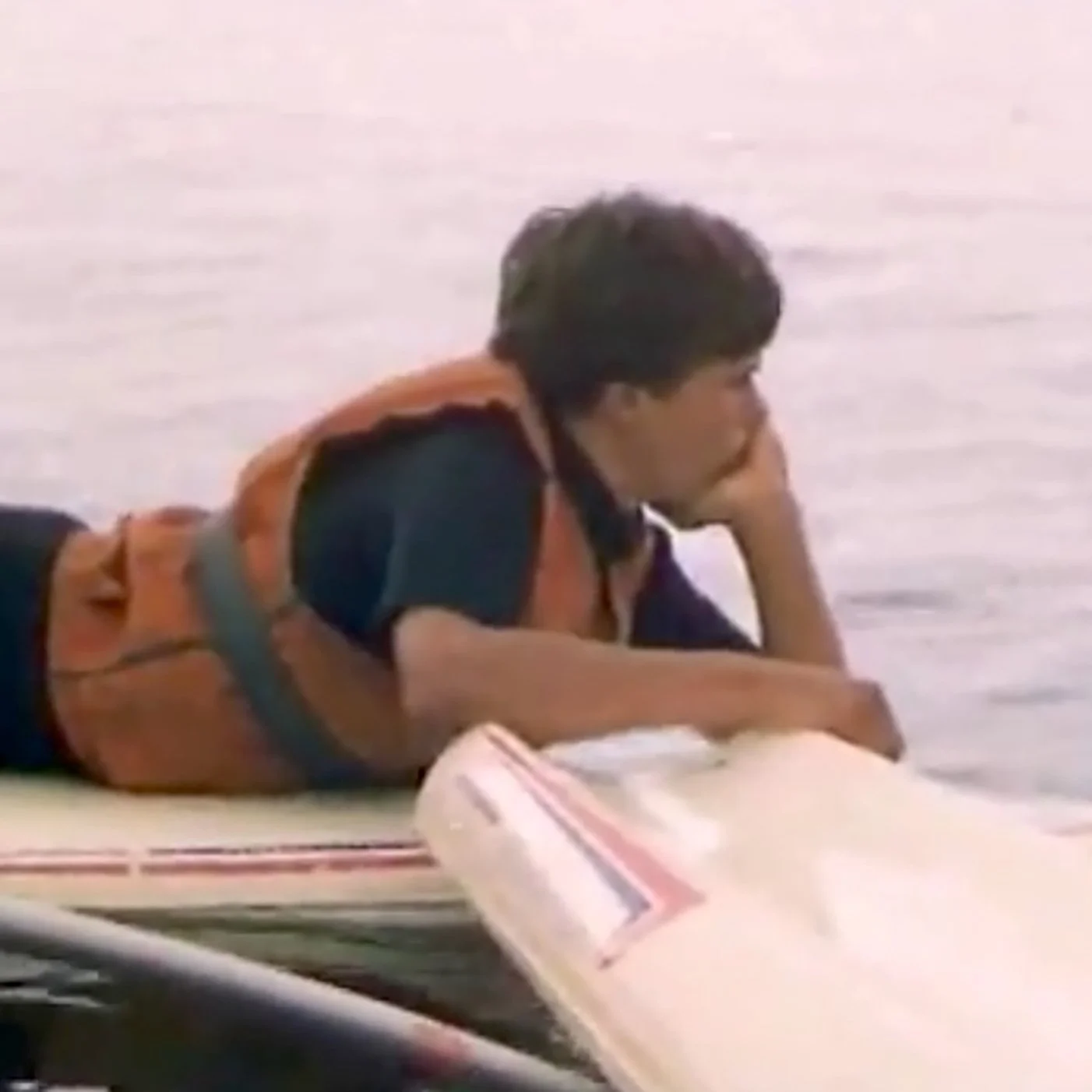 A young man lies on a boat with his head resting on his hand, wearing an orange life jacket over a dark shirt, in a body of water.
