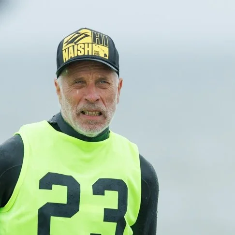An older man with a gray beard and mustache, wearing a black and yellow cap and a black athletic shirt under a neon green vest with the number 23. He has a concerned or confused expression on his face, and the background is a gray, overcast sky.