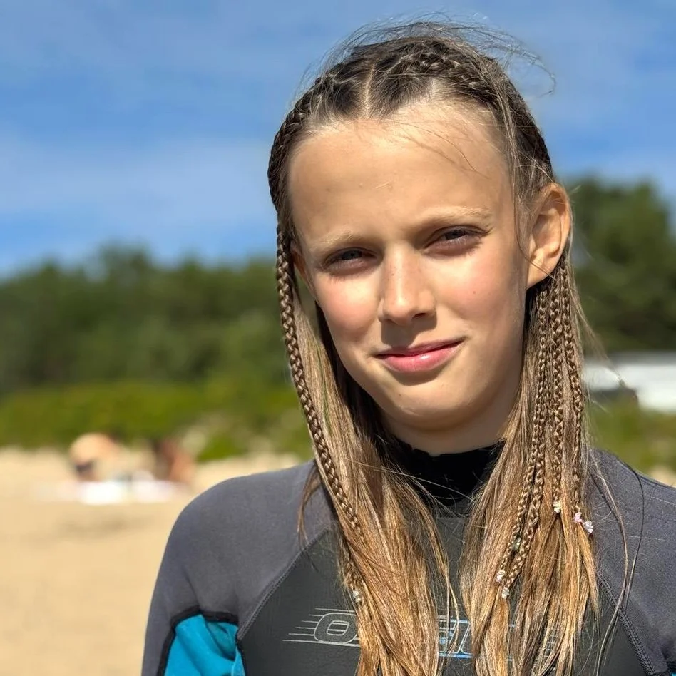 A girl with long hair and small braids, standing outdoors on a sunny day, with a beach and trees in the background.