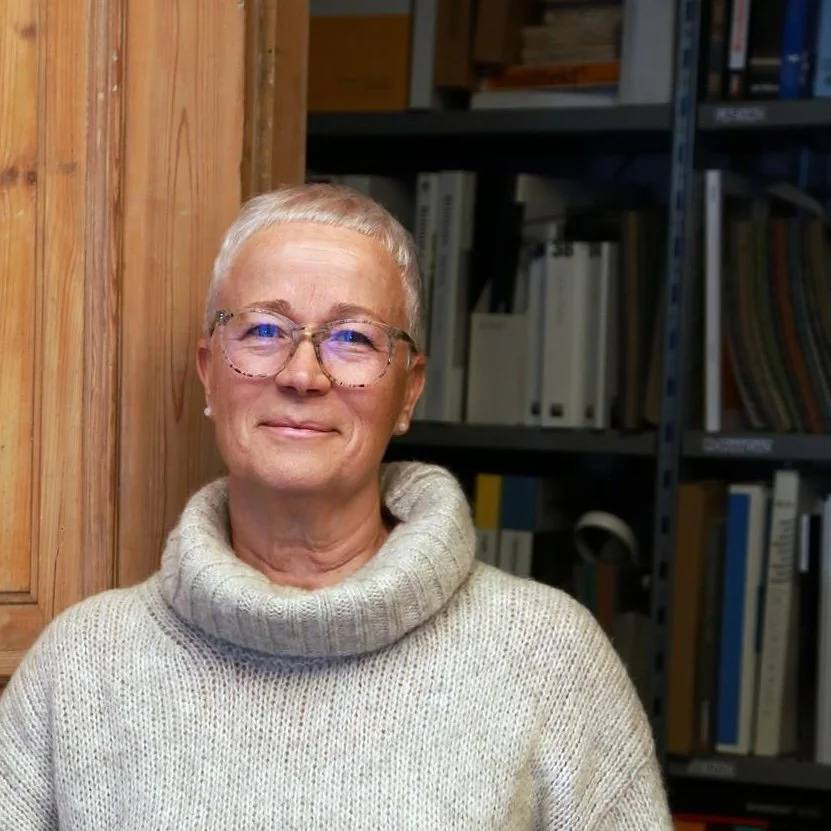 A woman with short gray hair, wearing glasses and a cream-colored turtleneck sweater, standing indoors against a wooden wall and a bookshelf filled with books.