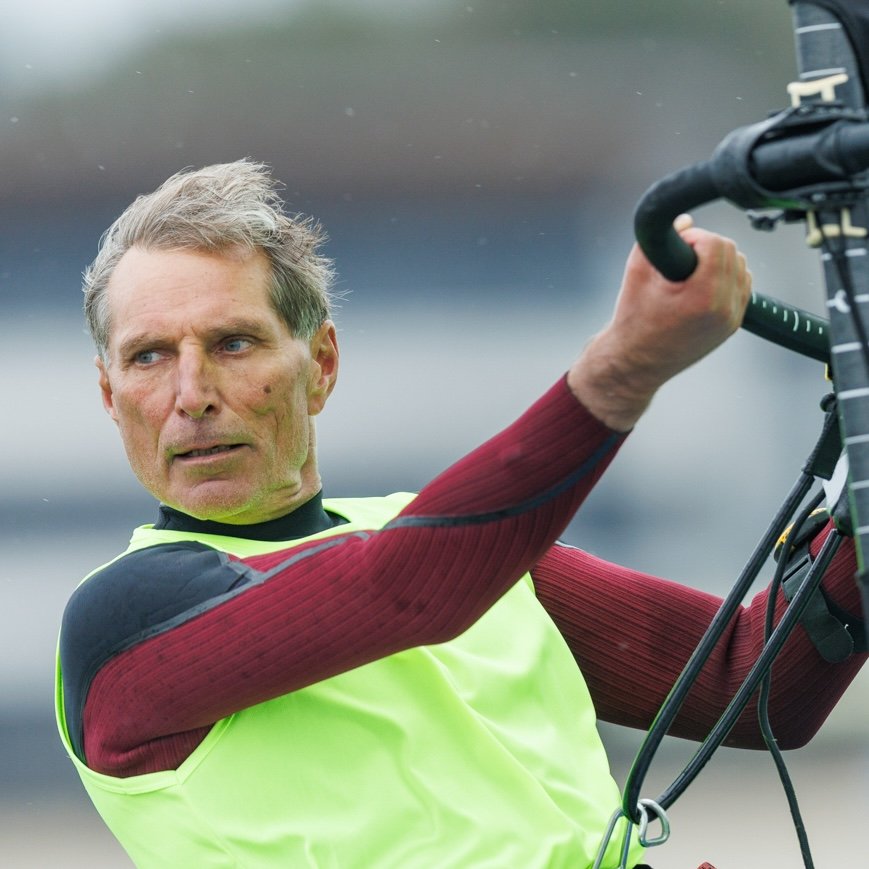 A man with gray hair wearing a neon yellow safety vest, long-sleeved red and black shirt, vision training goggles, and holding onto a control stick, standing outdoors on a cloudy day.