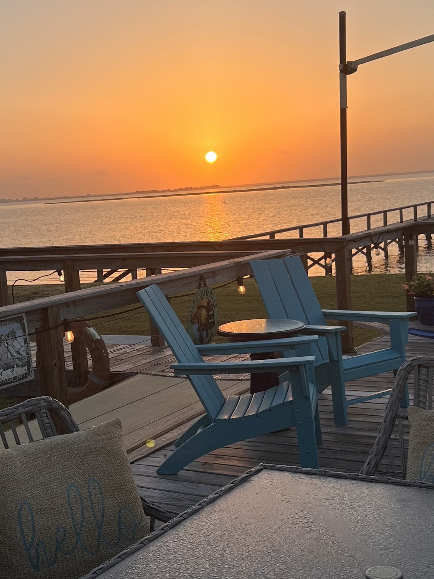 A lakeside deck during sunset with blue Adirondack chairs and a small round table, overlooking calm water and a colorful sky.