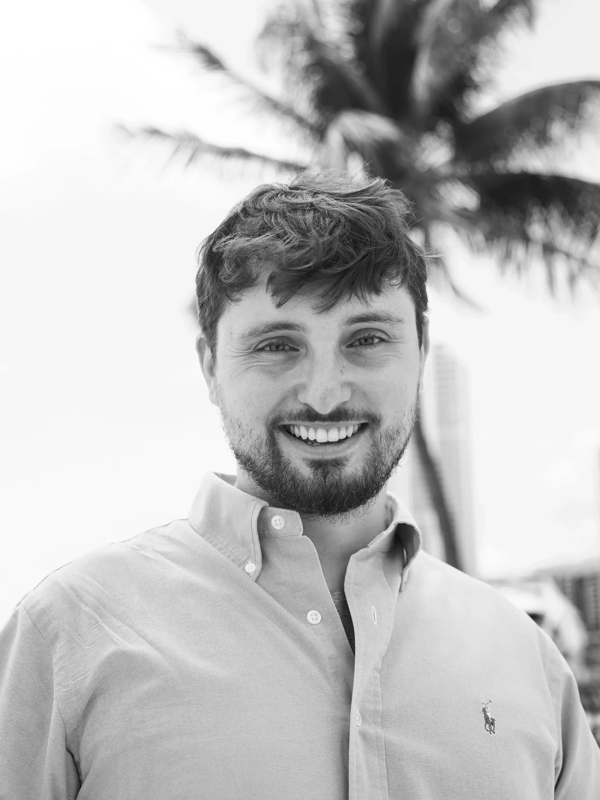 A smiling young man with light hair and a beard, wearing a light-colored collared shirt with a small embroidered horse and rider logo, standing outdoors in front of a palm tree.