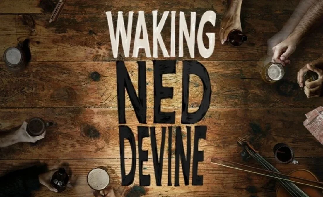 People sitting around a wooden table holding drinks, with the phrase 'Waking NED Devine' written in large letters on the table.