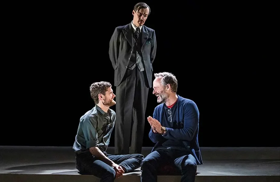 Three men on stage, one standing and two sitting, engaged in a conversation or performance, with a dark background.