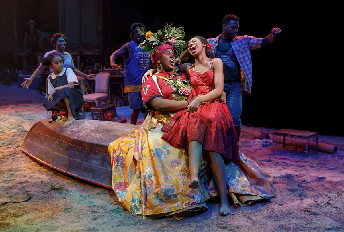 Theater scene with five actors celebrating on a stage that looks like sand, with a boat and chairs in the background. Two women are sitting on the boat, laughing and holding each other, while three others stand behind them, also smiling and enjoying the moment. The scene is lively and colorful.