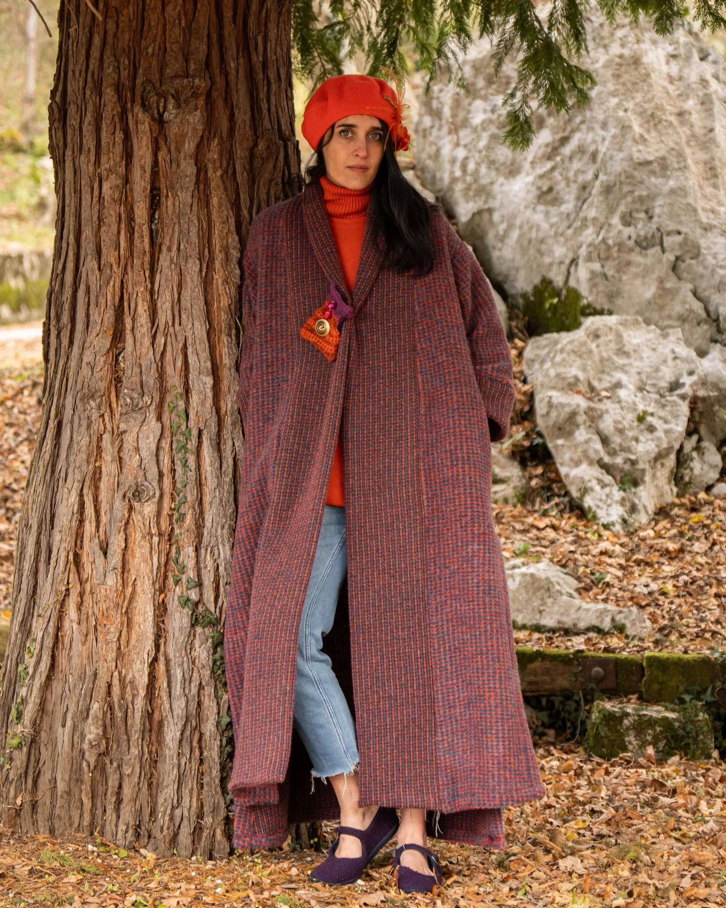Una donna indossa un cappotto lungo a motivi a scacchi sul tronco di un albero in un paesaggio autunnale con foglie e rocce.