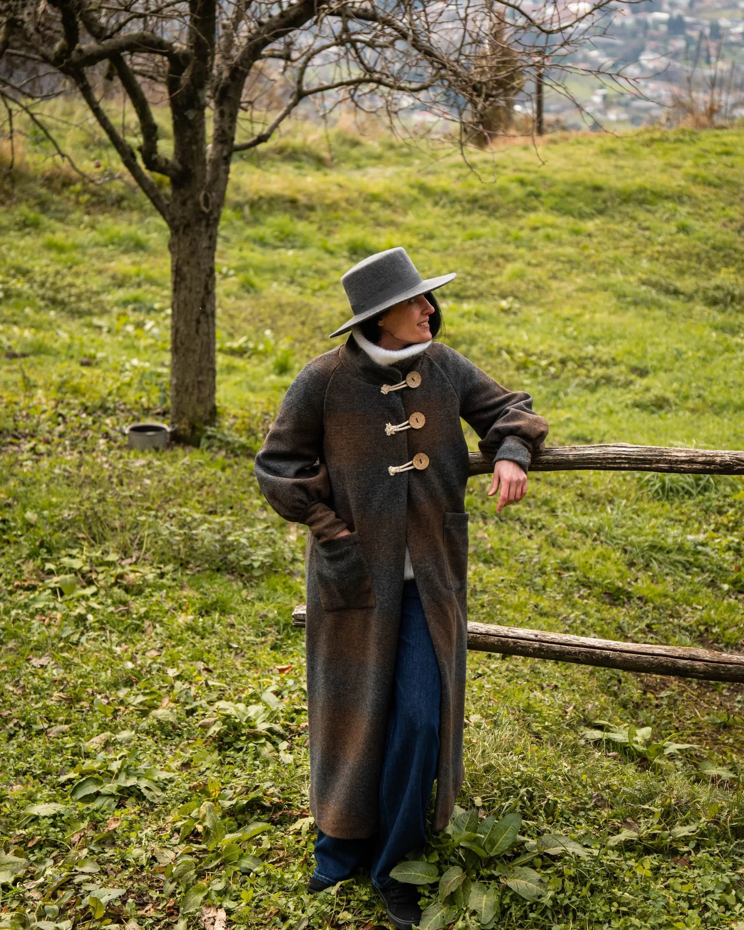 Una donna indossa un cappotto lungo e un cappello grigio mentre si appoggia a una staccionata di legno in un paesaggio verde con alberi privi di foglie e un cielo nuvoloso.