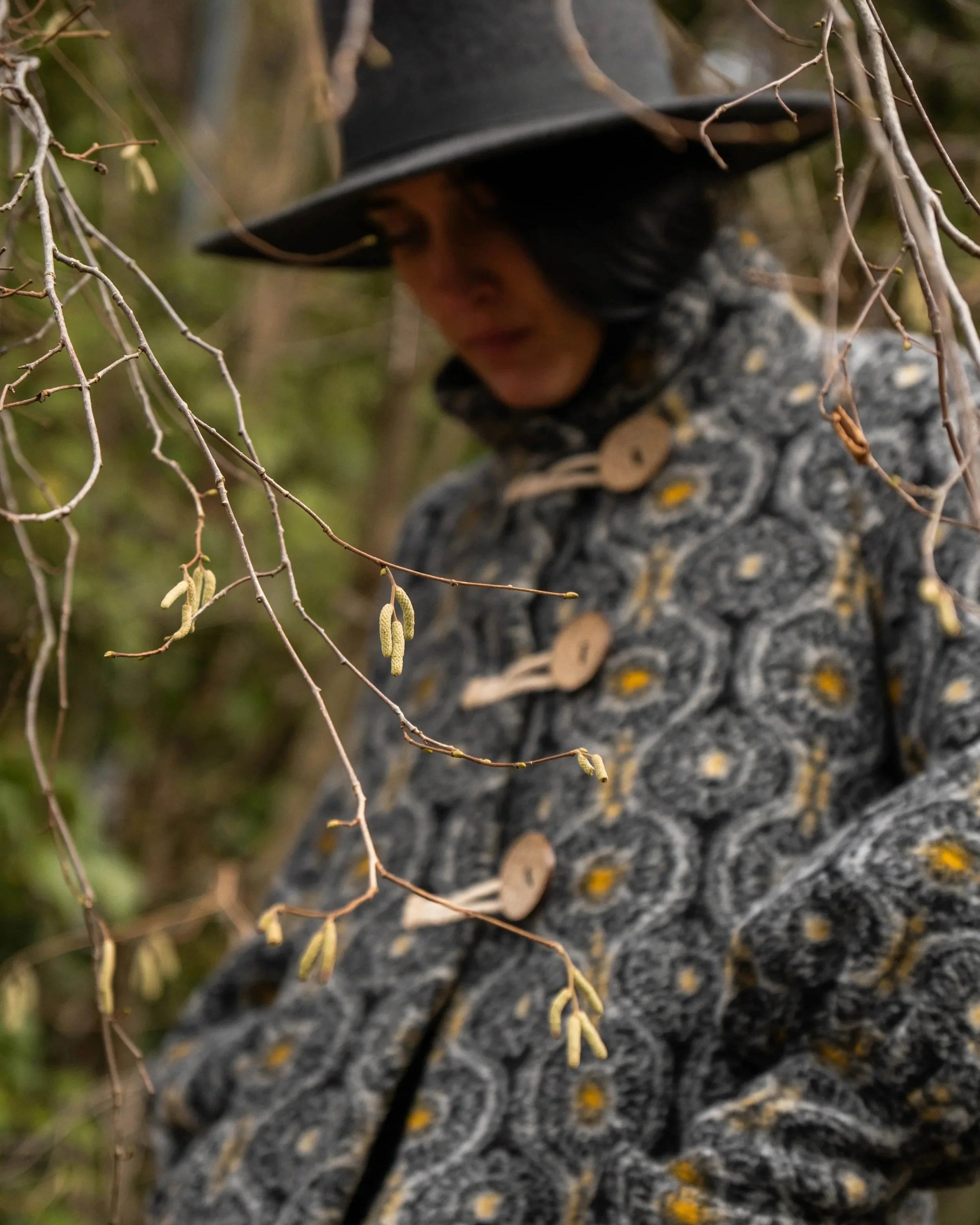 Donna indossando un cappello nero e un cappotto con motivo geometrico mentre si trova tra i rami di un albero.