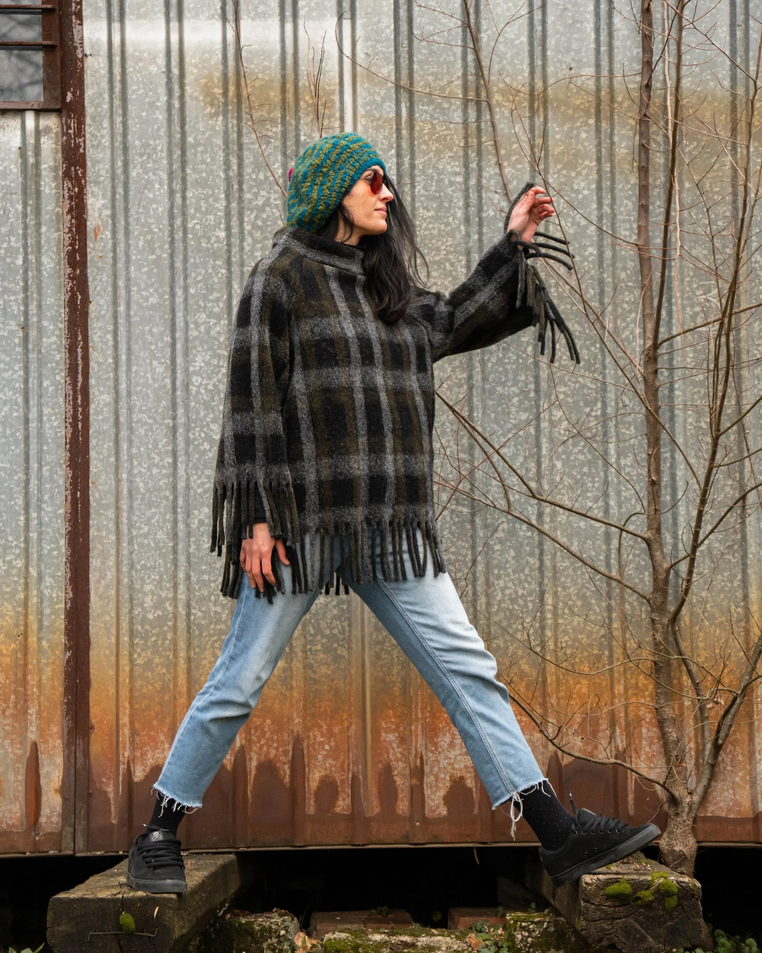 Una donna in stile grunge, indossa un cappello blu e verde, occhiali rossi, un cappotto scozzese, jeans, scarpe nere e sete nere con pois, mentre si appoggia a un albero senza foglie davanti a una recinzione in metallo arrugginita.