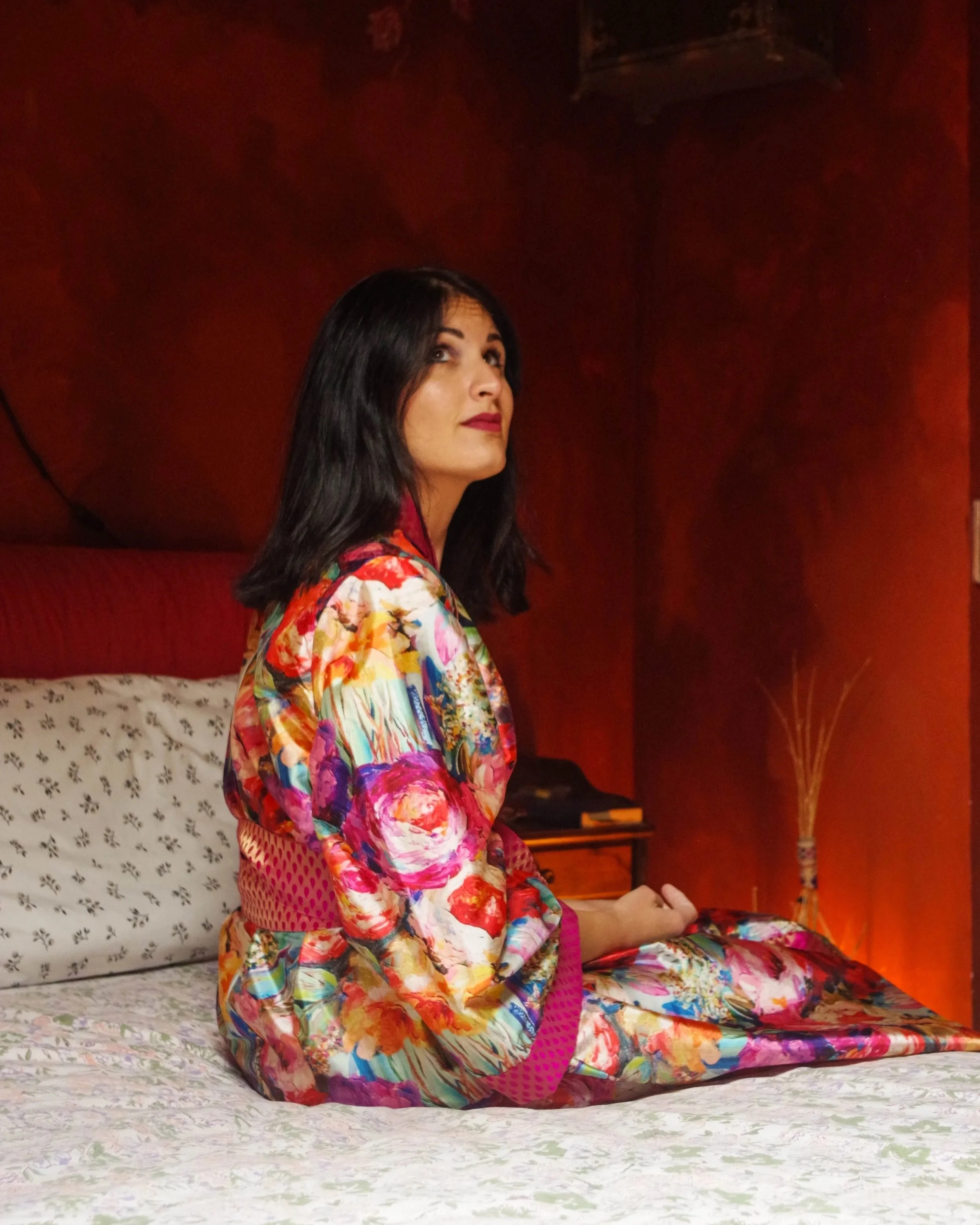 Una donna con capelli neri e trucco scuro, seduta sul letto in una stanza con pareti di legno scuro, indossa un kimono colorato con motivi floreali.