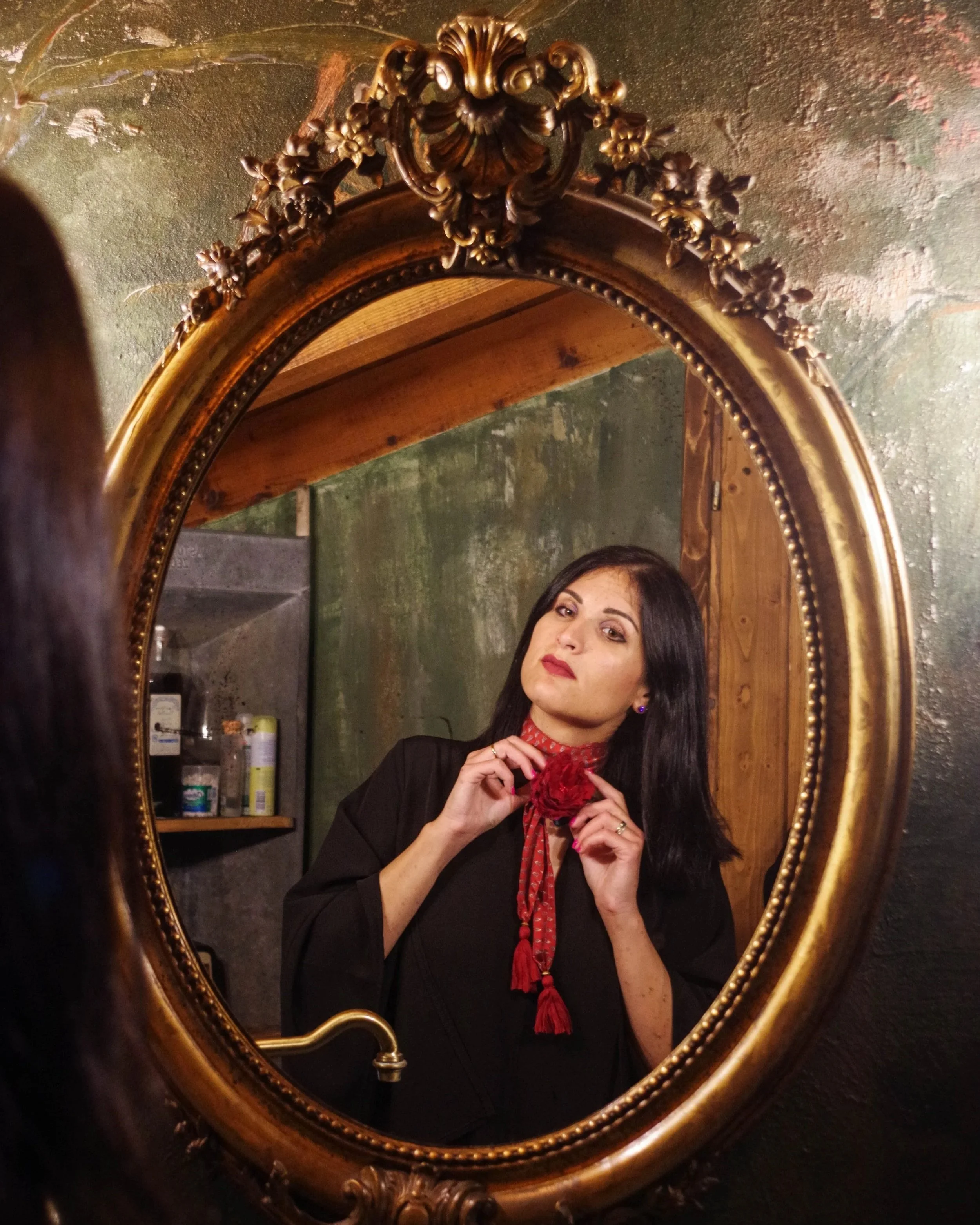 Ragazza con capelli scuri, trucco marcato, che si sistemando un foulard rosso intorno al collo, vista attraverso uno specchio dorato vintage con decorazioni ornate.