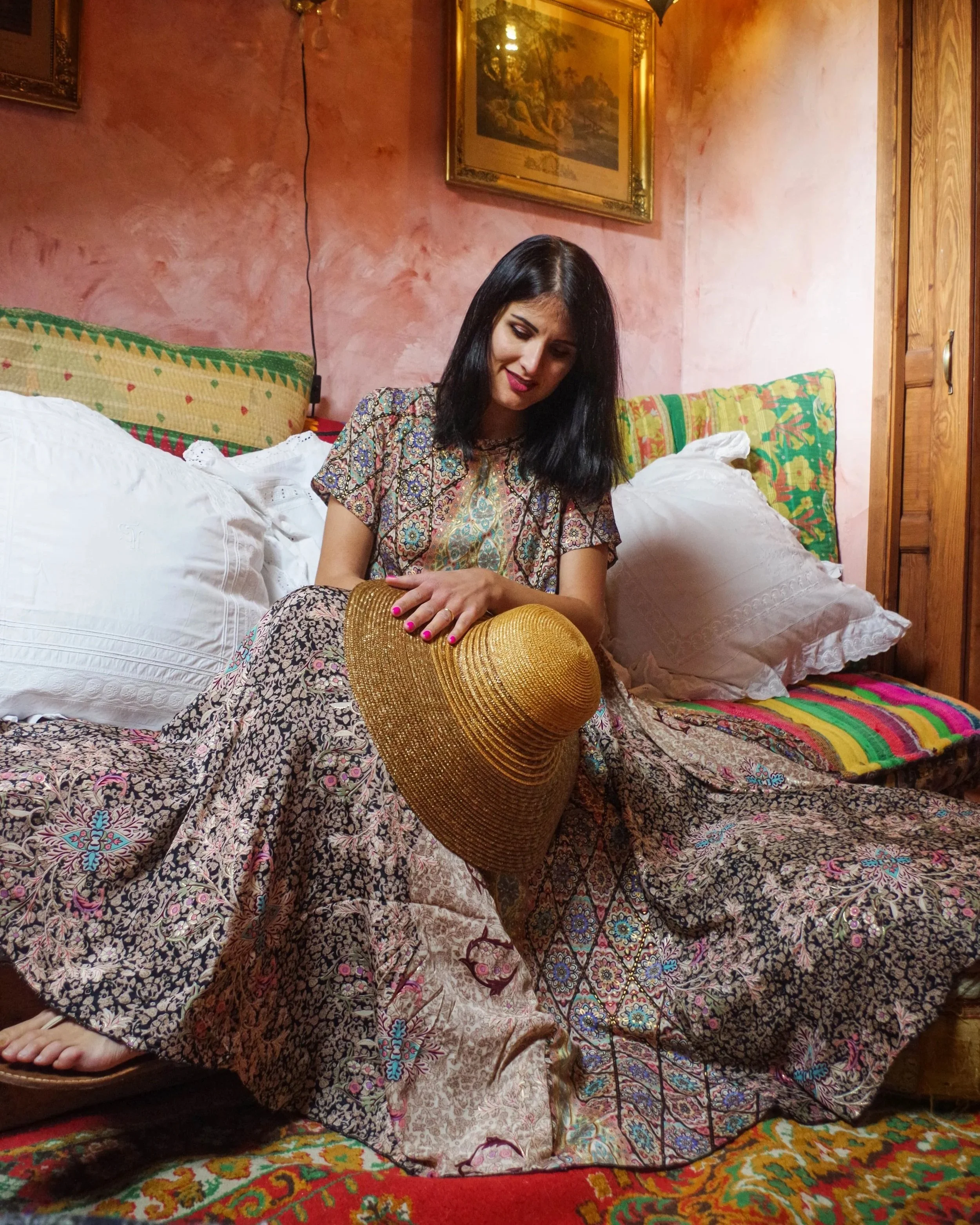 Una donna seduta su un letto colorato con cuscini e copriletto, che tiene un cappello di paglia in grembo, in una stanza con pareti rosa e quadri appesi.