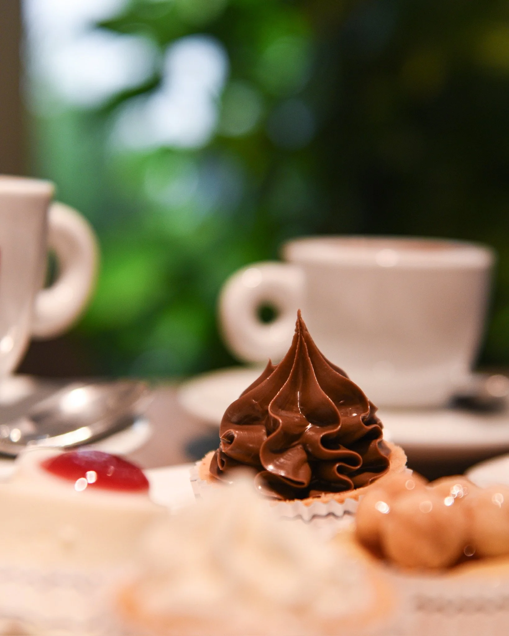 Dolce al cioccolato con decorazione a pizzo di cioccolato, accompagnato da altri dolci e tazze sul tavolo.