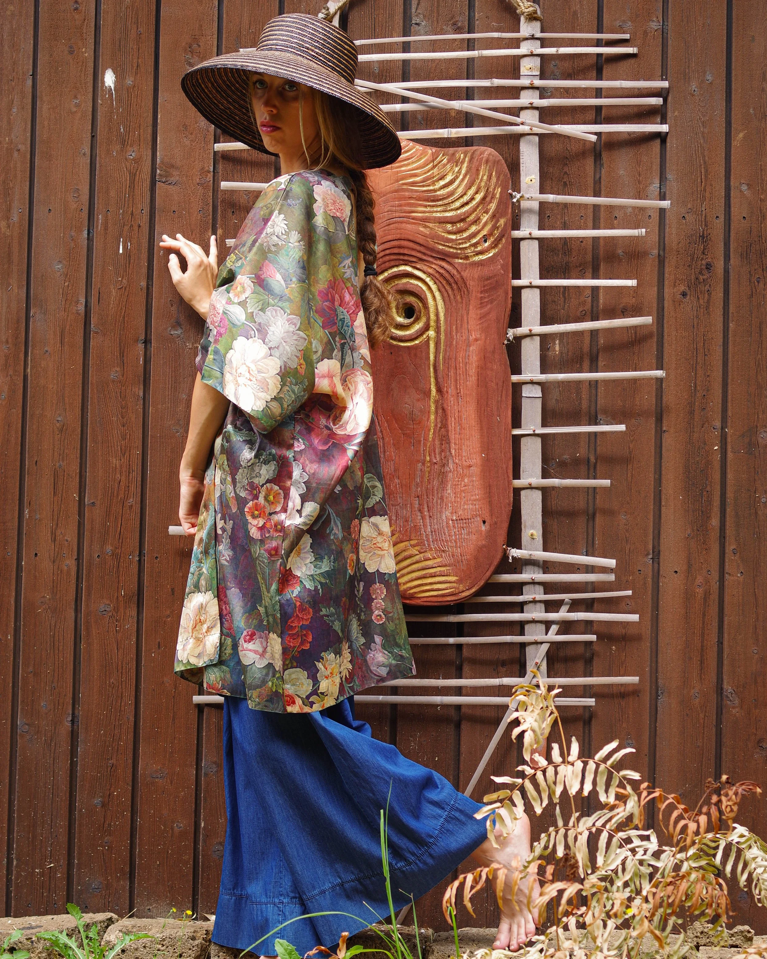 Una donna indossa un cappello a larghe tese, un kimono floreale e un paio di pantaloni blu, si trova davanti a una parete di legno con un'installazione artistica in legno