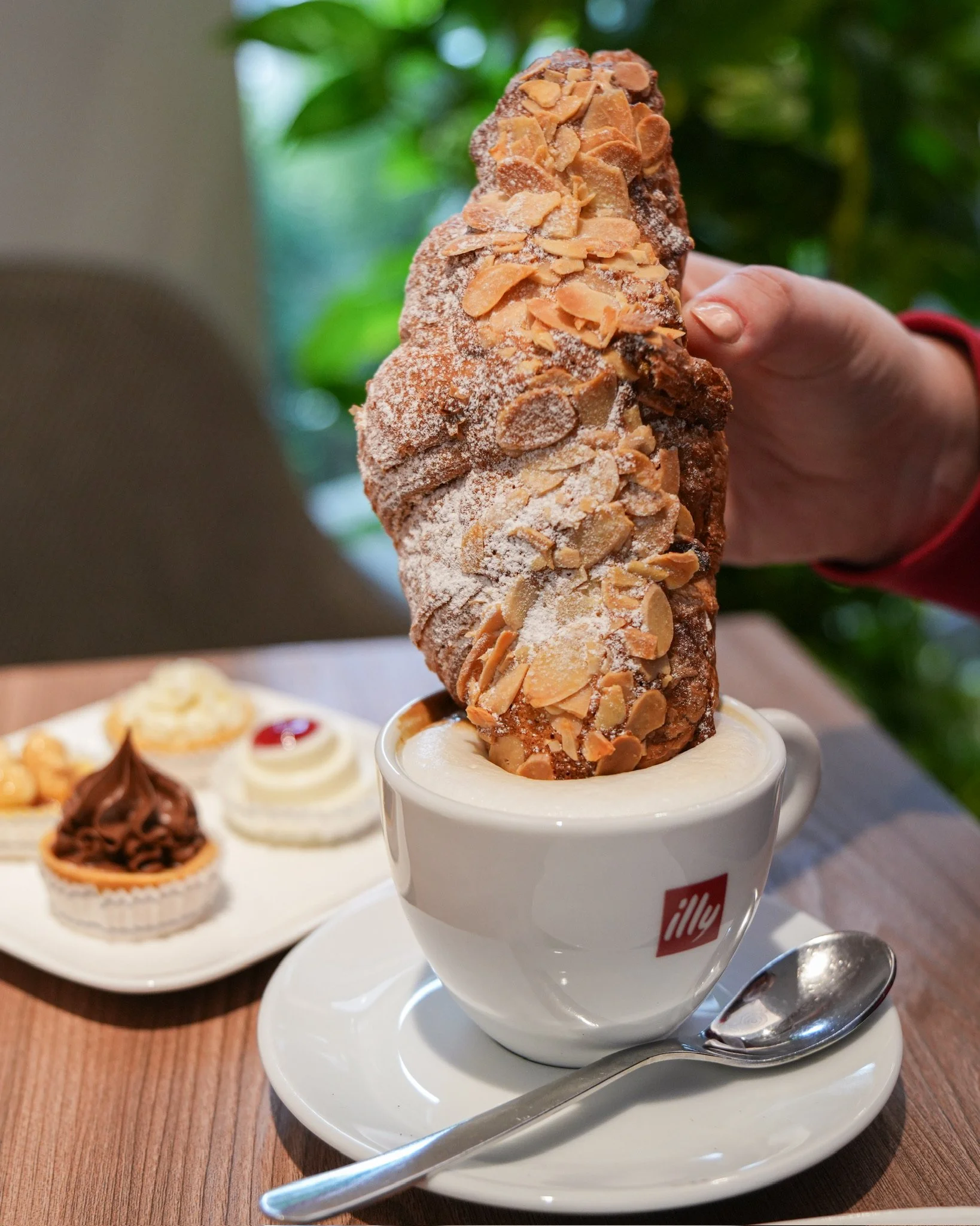 Una persona tiene in mano una grande brioche con mandorle e zucchero, immersa in una tazza di latte o cappuccino con il logo illy, sullo sfondo ci sono altri dolci, come una ciambella con cioccolato e una crema con ciliegina.