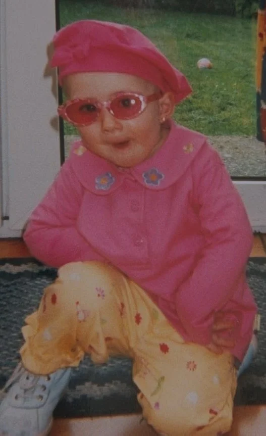 Une petite fille souriante portant des lunettes de soleil roses, un chapeau rose, une veste rose avec des broderies bleues, un pantalon jaune avec des motifs rouges, assise à l'intérieur près d'une porte ou fenêtre, avec un jardin en arrière-plan.
