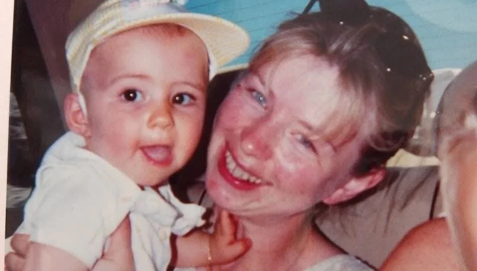 Une jeune femme avec une orchidée dans les cheveux sourit en tenant une jeune fille portant un chapeau blanc et une chemise blanche, tous deux en plein air.