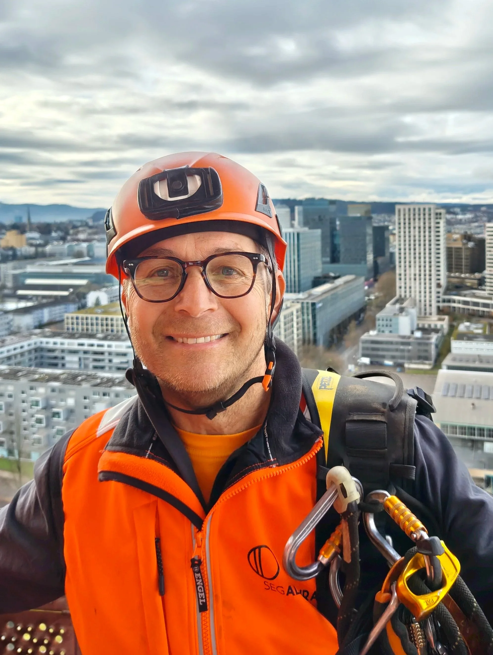 Bruno Lochmann in orange Sicherheitskleidung mit Helm und Kletterausrüstung auf einem Hochhaus-Baustandort in einer Stadt mit modernen Gebäuden, bewölktem Himmel.