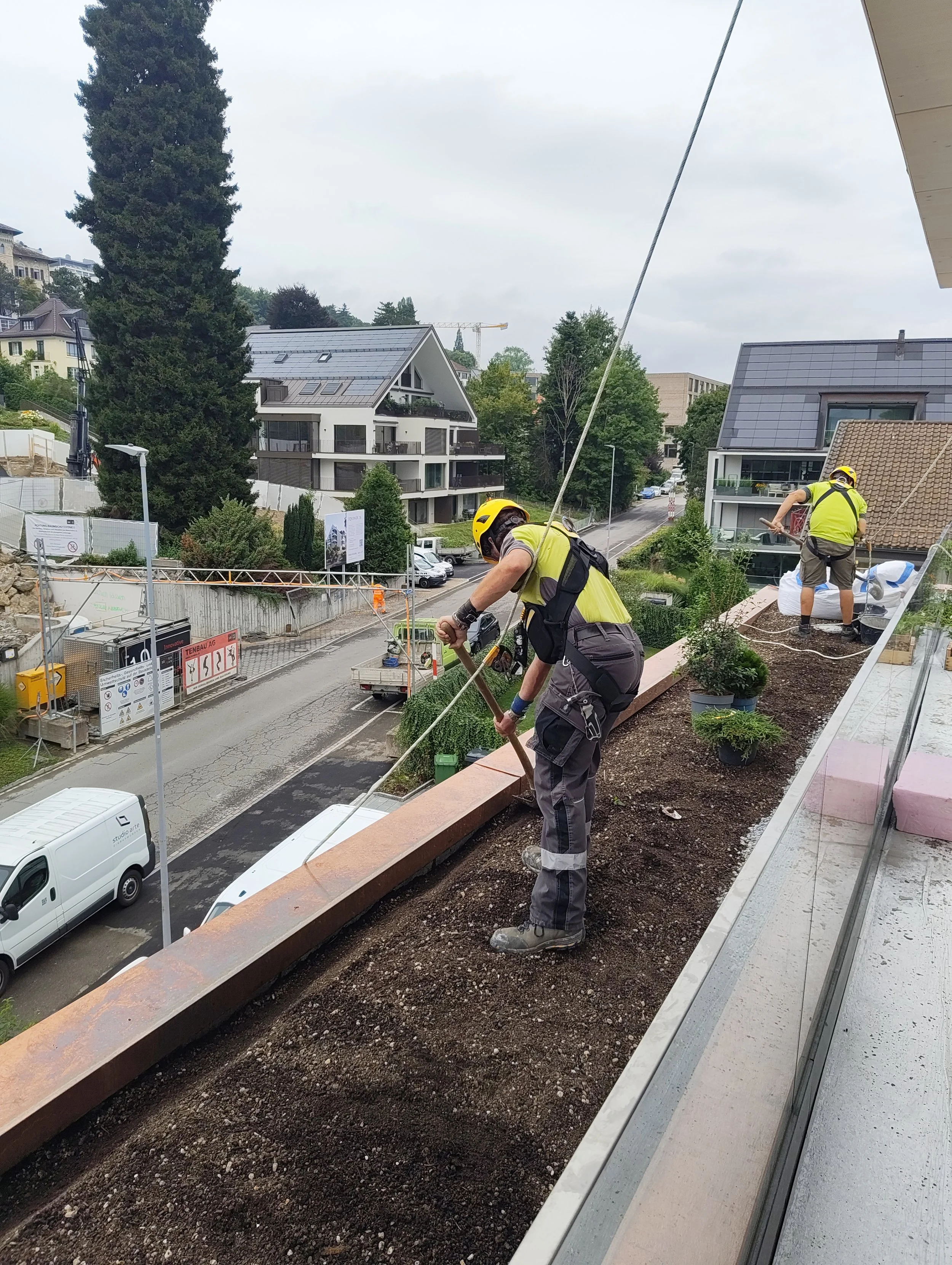 Zwei Bauarbeiter, gesichert durch temporäre Absturzsicherungen, pflanzen Boden auf einer Terrasse mit Blick auf eine Straße und moderne Wohnhäuser.