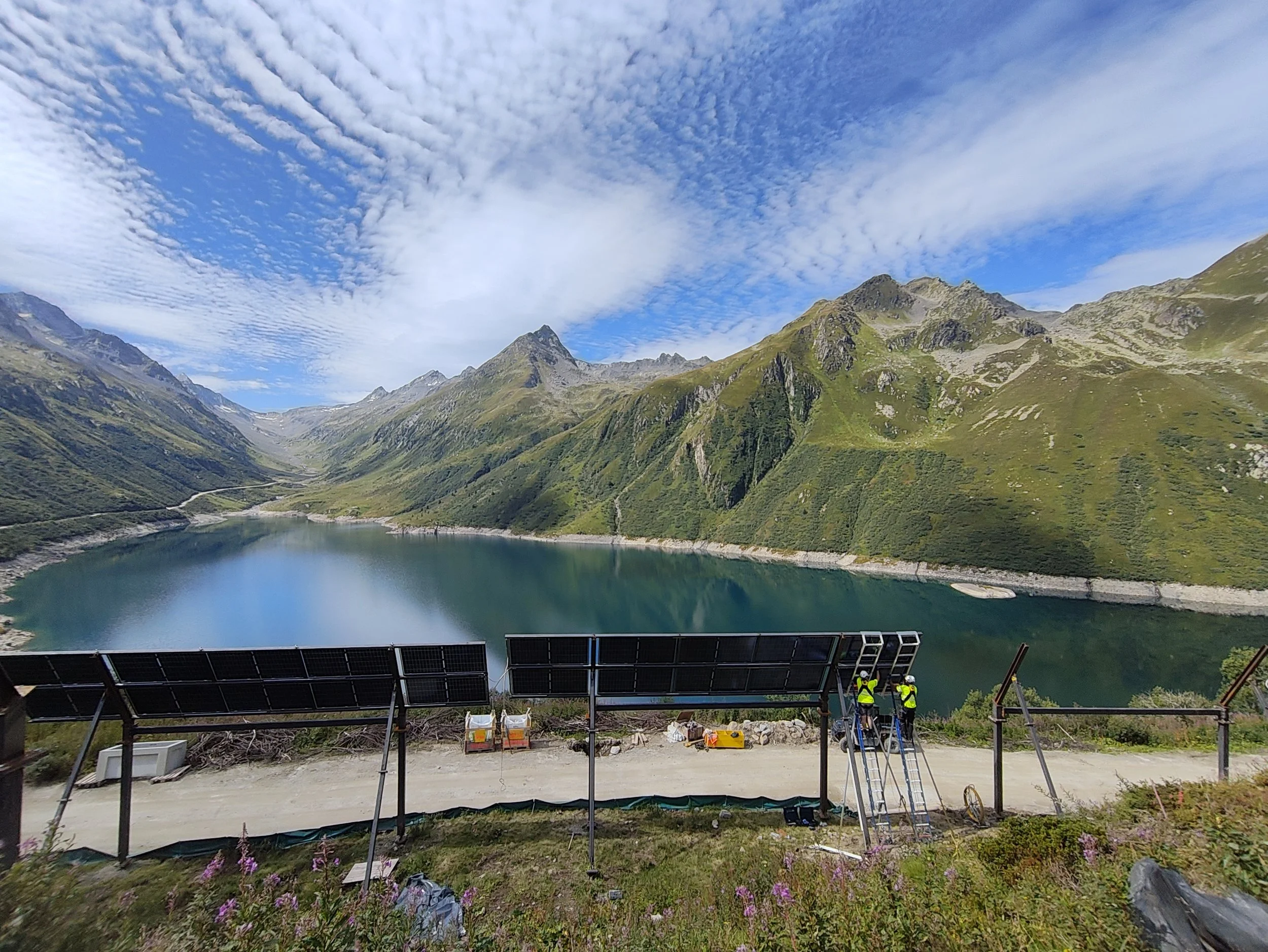 Blick auf eine Berglandschaft mit einem See im Vordergrund und einem blauen Himmel mit Wolken, im Vordergrund mehrere Personen beim Arbeiten an Solaranlagen.