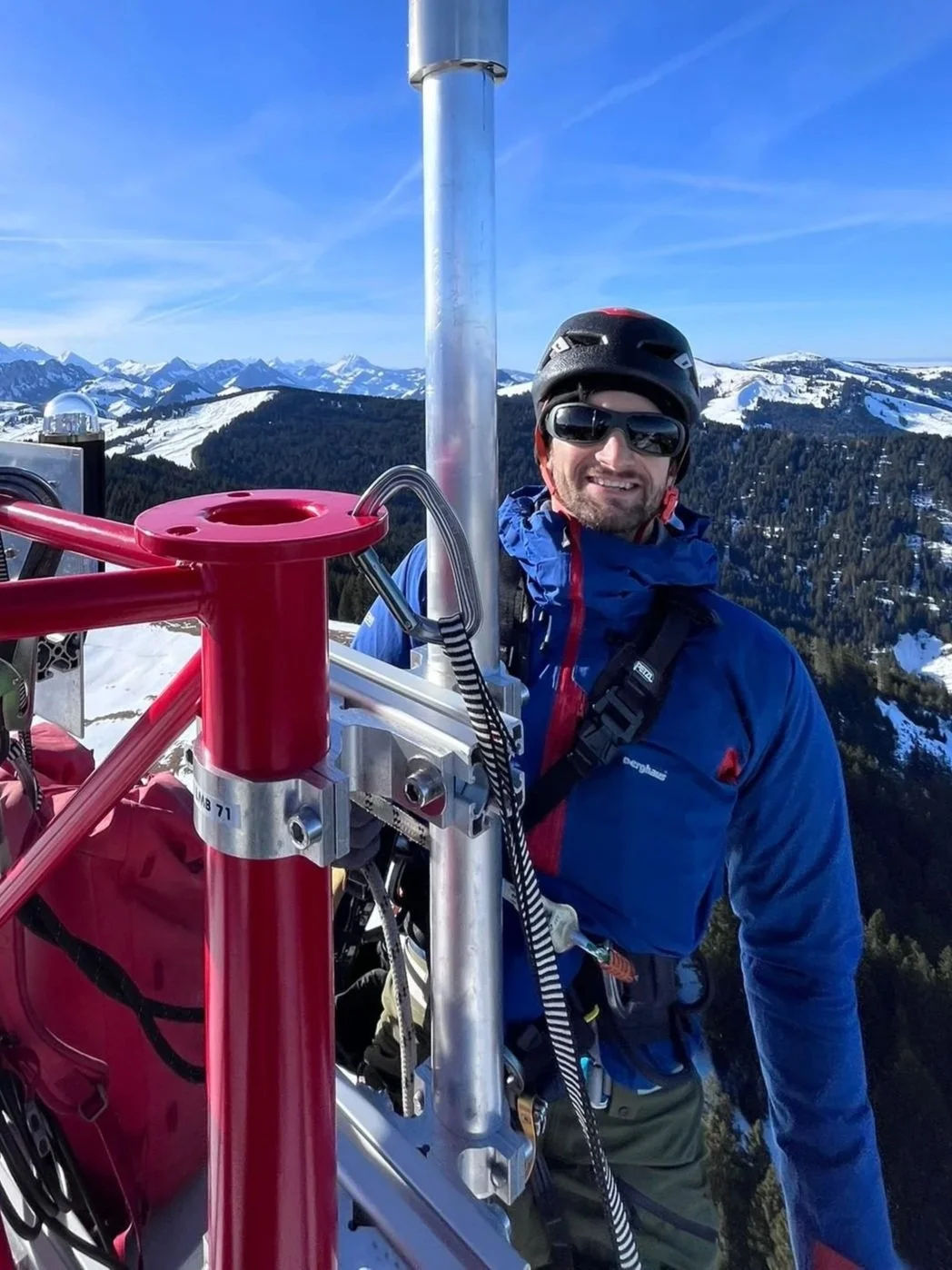 Stefan Birrer in Bergsteigerkleidung, mit Helm und Sonnenbrille, steht auf einer Messanlage mit Schneelandschaft und Bergen im Hintergrund, neben einer technischen Vorrichtung.