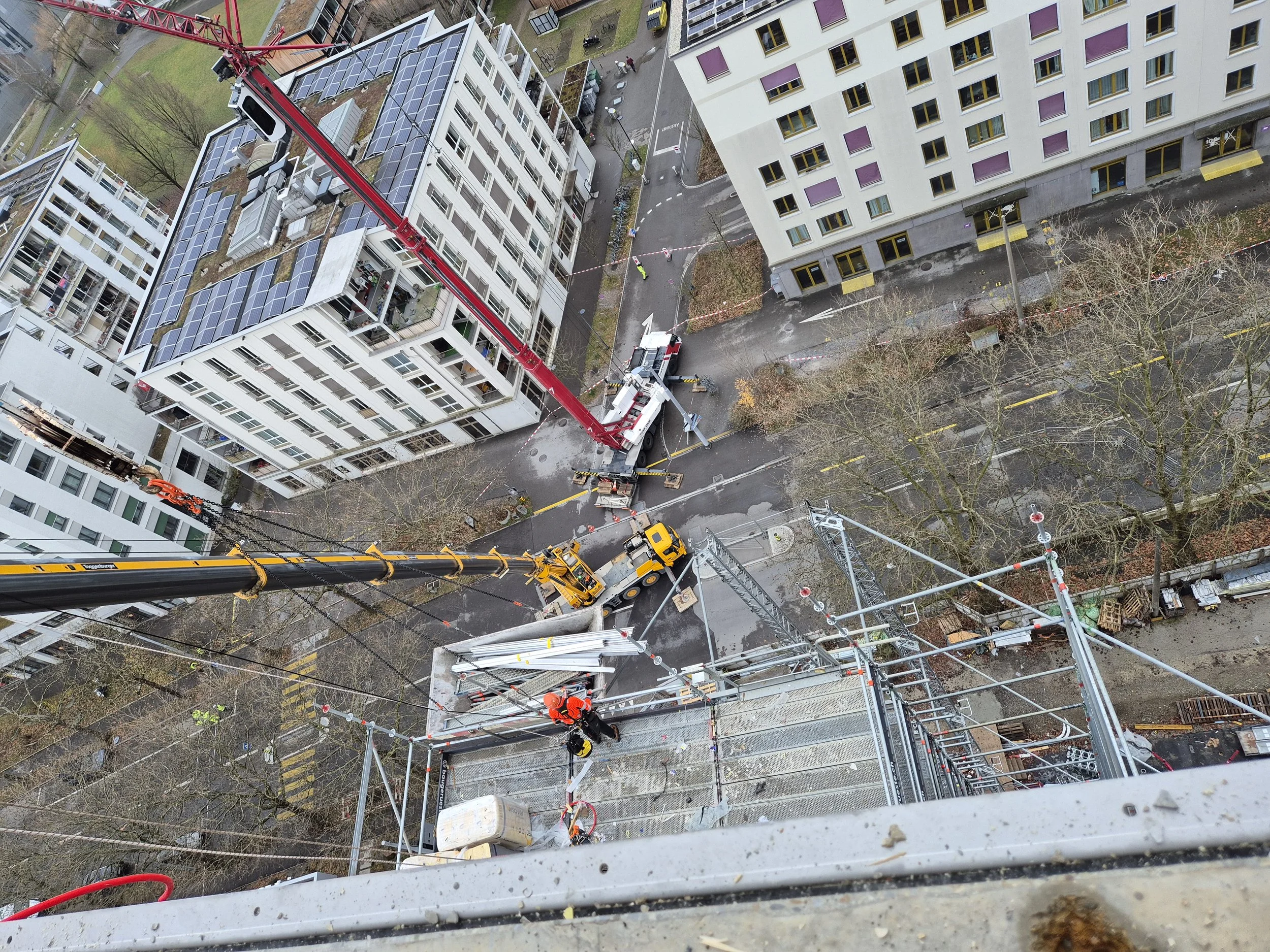 Ein Blick von oben auf eine Baustelle in einer Stadt mit einer hohen Kran, Gebäuden, Straßen und einigen Bäumen, die im Herbstlaub stehen.