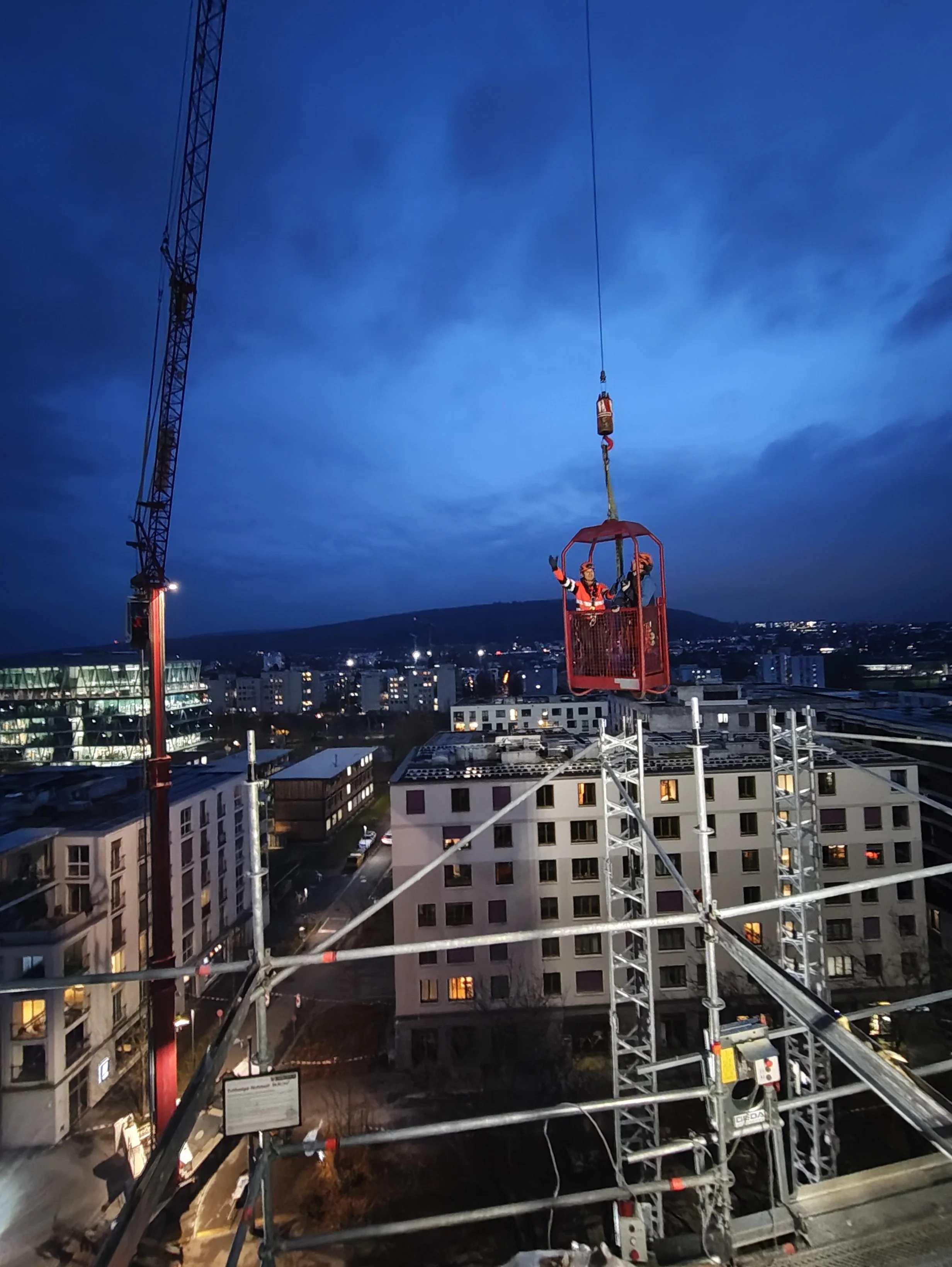 Auf einer Baustelle in der Abenddämmerung sind Gerüste und Kranstrukturen zu sehen. Zwei Arbeiter in Schutzkleidung und Helmen stehen in einem Korb, der an einem Seil aufgehängt ist. Im Hintergrund sind Häuser und eine Stadtlandschaft bei twilight sichtbar.