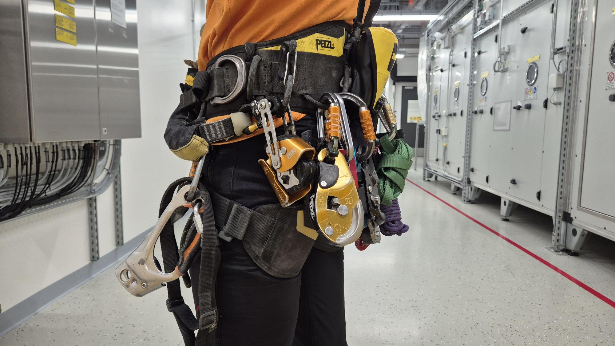 Person mit Kletterausrüstung in einem technischen Raum. Die Ausrüstung umfasst Karabiner, Seile und andere Kletterinstrumente, getragen um die Taille.