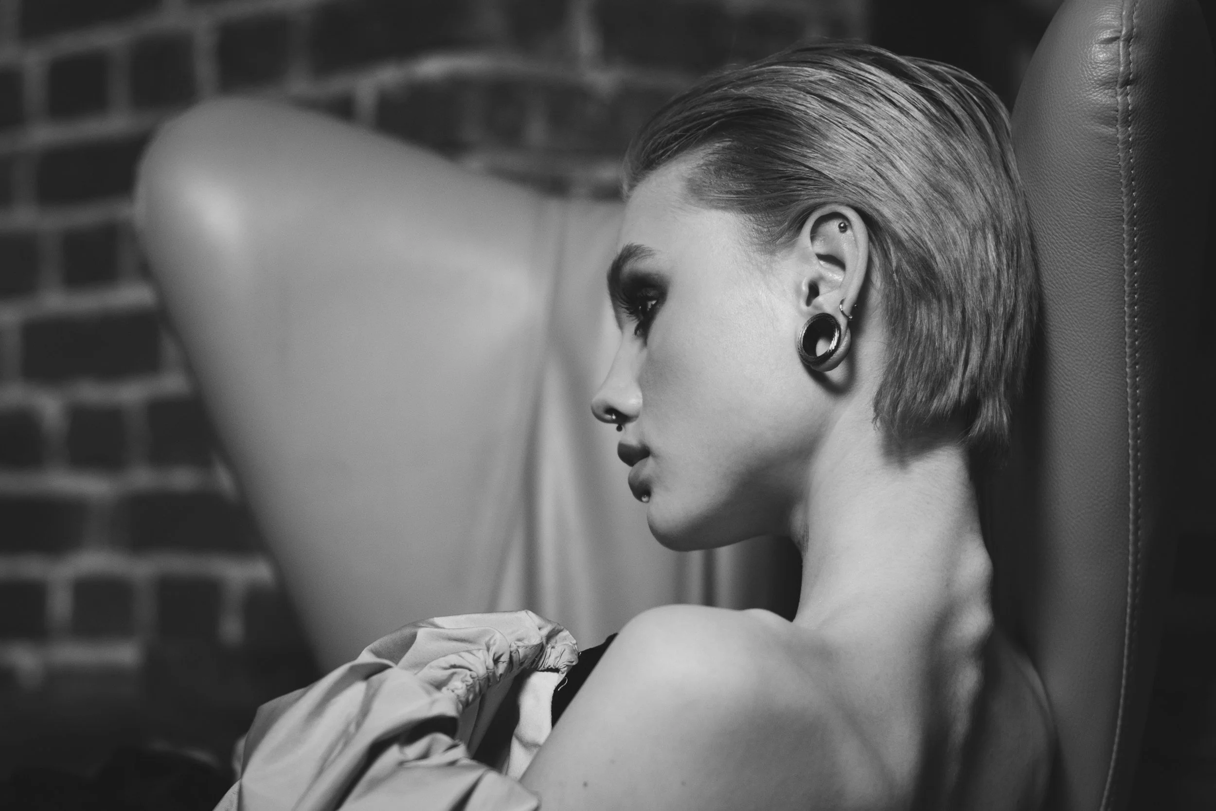 A black and white photo of a woman with short hair, wearing large stretched earrings, sitting against a brick wall at Carlisle castle piercing 

the piercing guy in Carlisle at immortal art studio