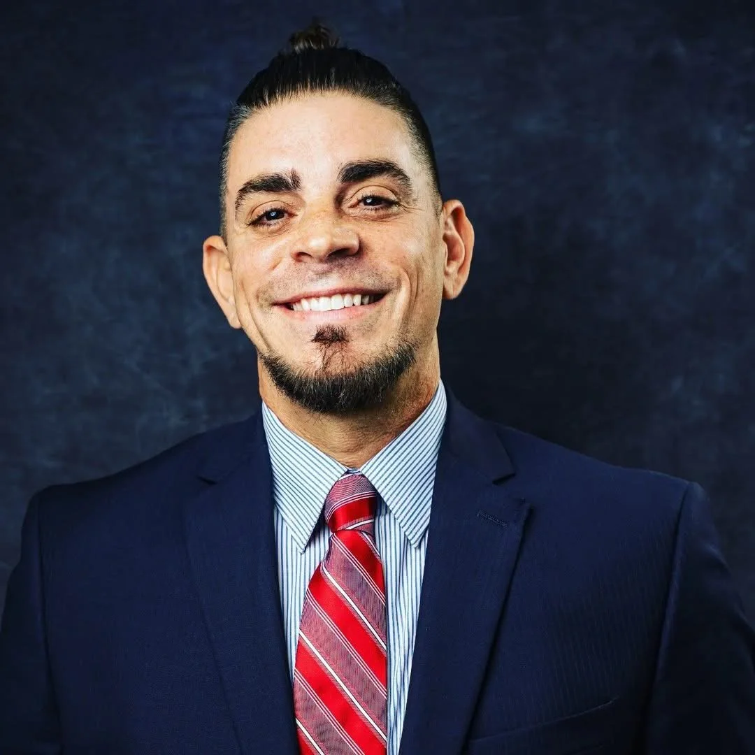 Headshot of a man wearing a navy suit, light blue collared shirt, and red striped tie, smiling against a dark background.