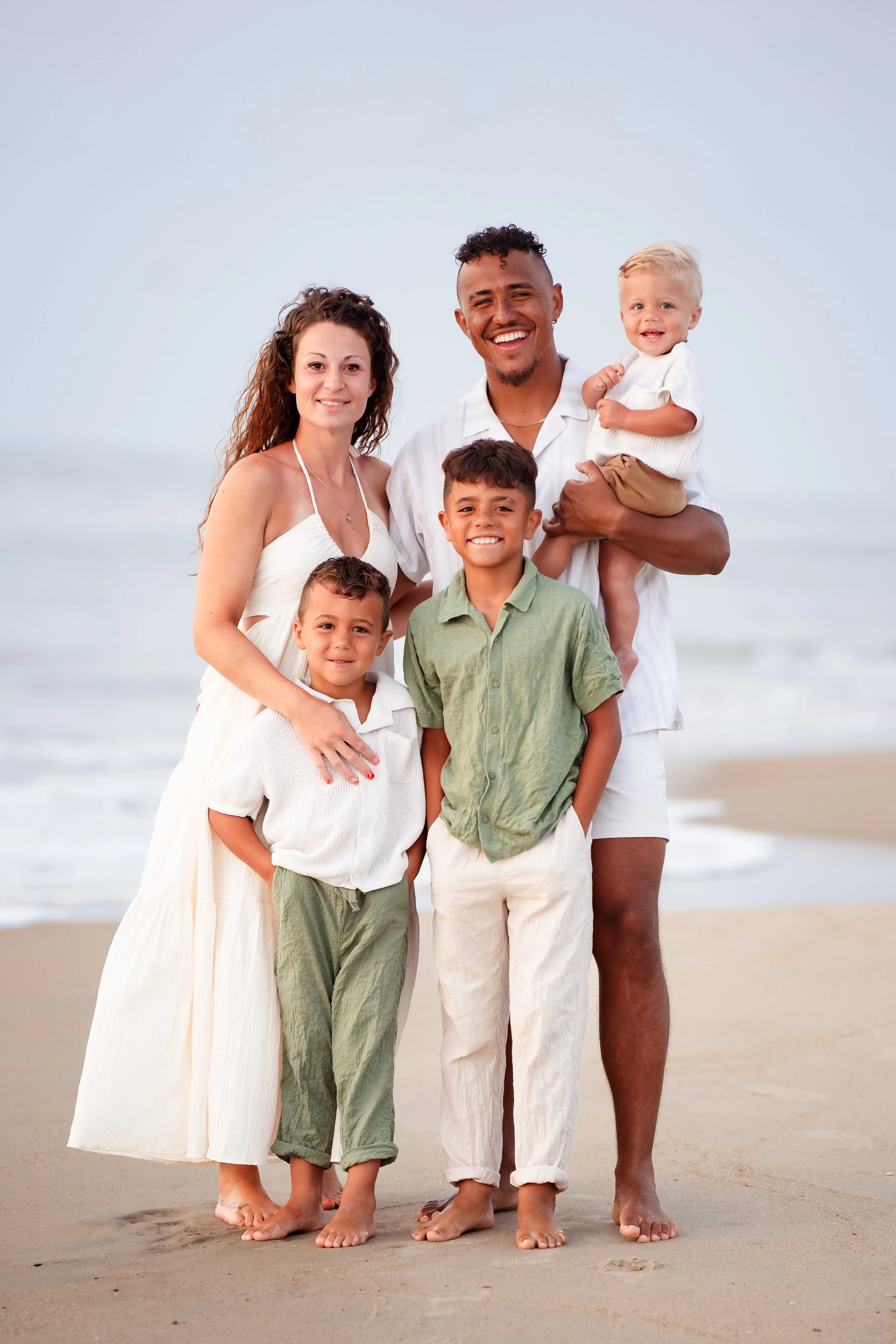 A diverse family of six standing on a sandy beach, smiling at the camera with the ocean in the background.