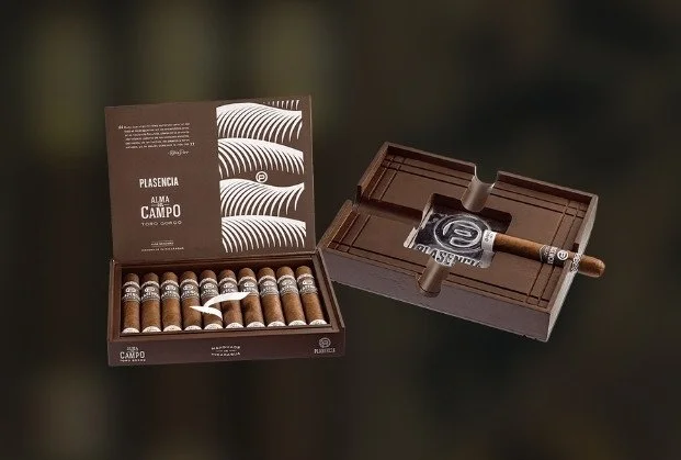 An open box of cigars with a booklet inside, and a lit cigar resting in an ashtray with a printed logo.