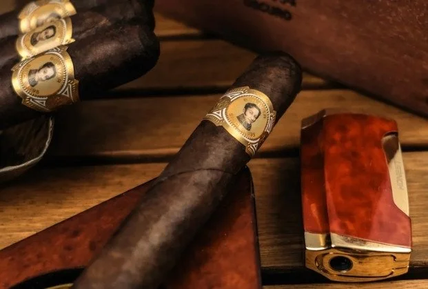 A cigar with a gold band featuring a man's portrait and a wooden cigar holder, along with a lighter on a wooden surface.