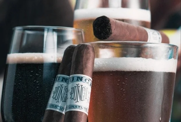 Two glasses of beer and two cigars resting on top of the glasses.