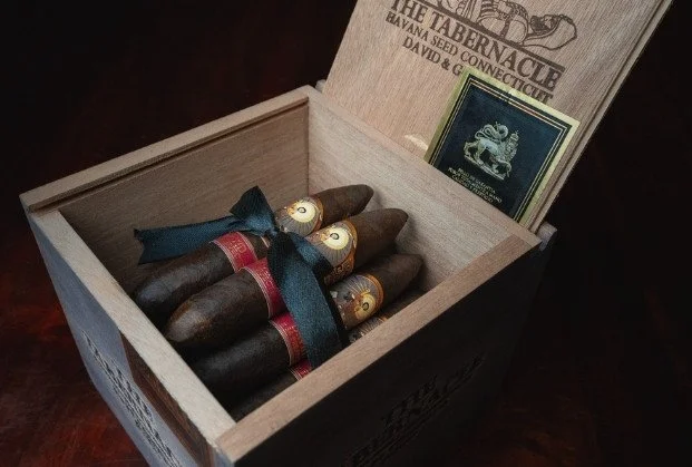 Open wooden box containing five cigars with red and gold bands, tied together with a blue ribbon, and a deck of tobacco cards.