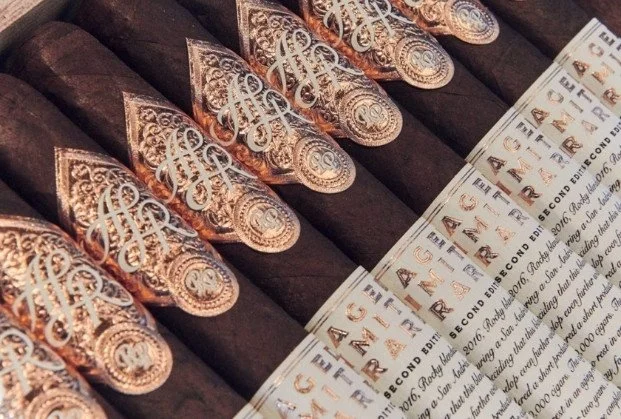Multiple partially lit cigars with decorative gold and silver banding, placed next to a printed label or certificate.