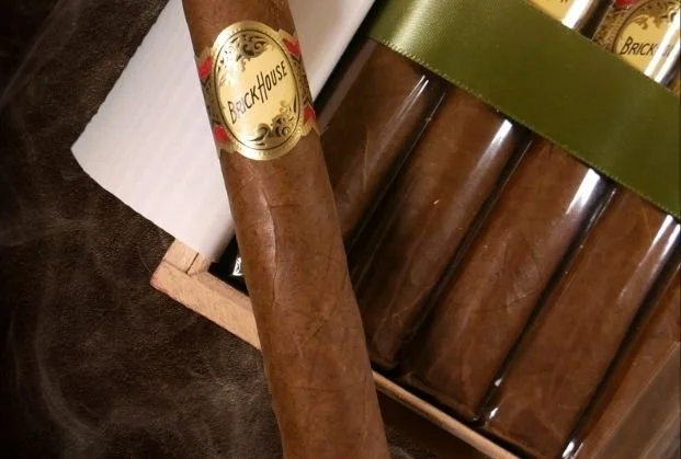 A close-up of a box of cigars, with one cigar outside the box marked 'Brick House'. The cigars inside the box are wrapped in plastic with a green band.