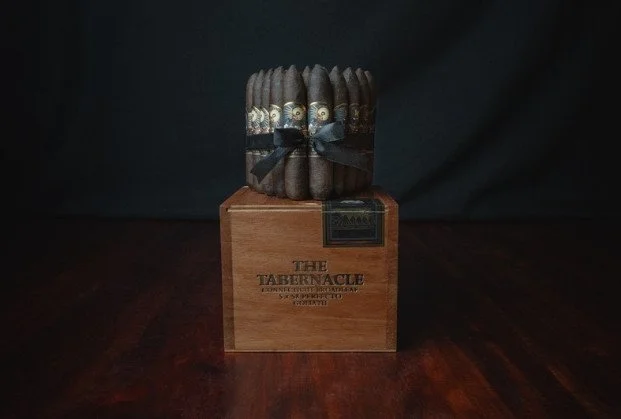 A bundle of cigars wrapped with a black ribbon, placed on a wooden box labeled 'The Tabernacle' with a dark background.