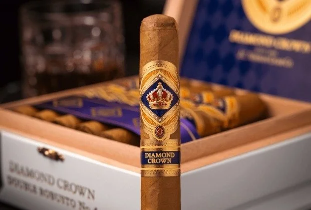A close-up of a Diamond Crown cigar standing upright with a blue and gold band, with a box of Diamond Crown cigars in the background.
