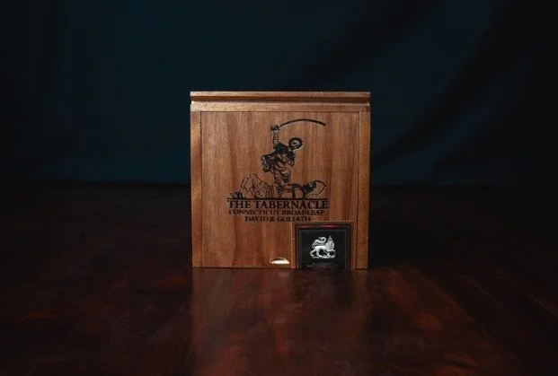 A wooden box with a carved figure of a fisherman holding a fishing rod, and a small framed photo or certificate attached to the front, placed on a dark wooden surface against a dark background.