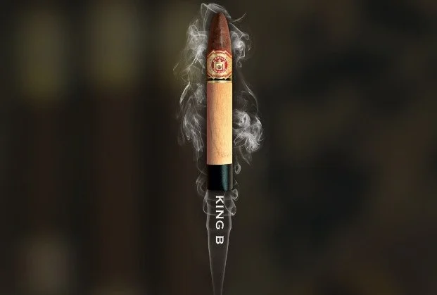 A cigar with smoke emanating from its tip, mounted on a black object labeled 'KING B' with a dark, blurred background.