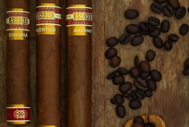Three cigars labeled Enigma, Actuijo, and Arcano next to scattered coffee beans on a wooden surface.