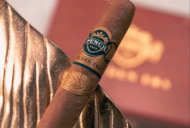 A Punch brand cigar resting on a silver metallic surface with a branded label and a gold band near the filter end, with a blurred box in the background.